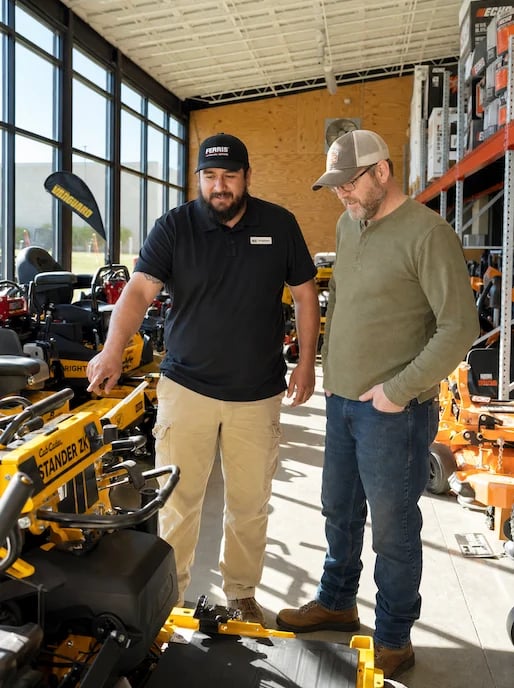 Two men in a warehouse showroom discussing yellow landscaping equipment. Large industrial windows and shelving visible in background.
