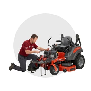Technician in red uniform kneeling beside a red and gray riding lawn mower for maintenance or inspection