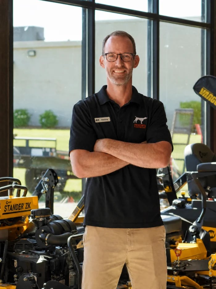 Professional man in black polo shirt standing with arms crossed in front of yellow heavy machinery, posing by large windows